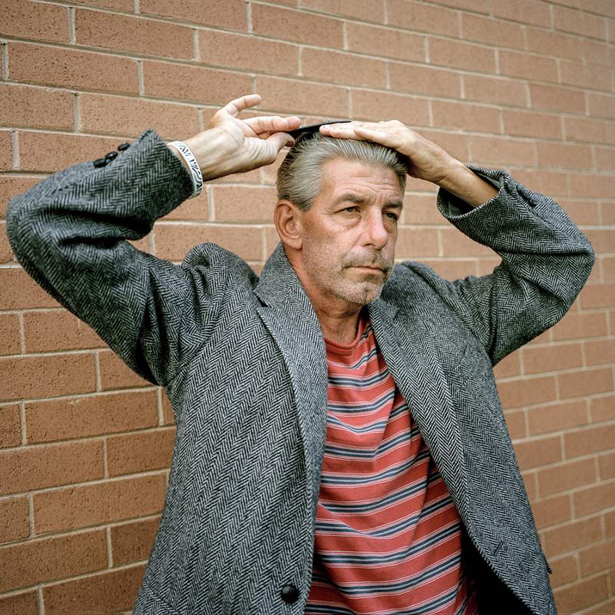 An older man, standing by a wall, combing his hair