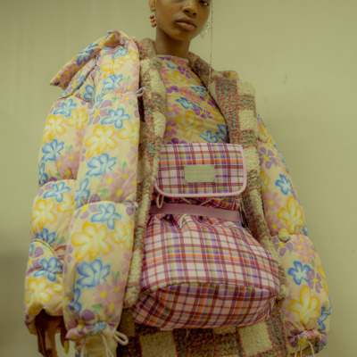 A girl with shaved head wears a floral puffer over a pink and white check set and matching floral top. She also wears a plaid pink backpack but on the front.