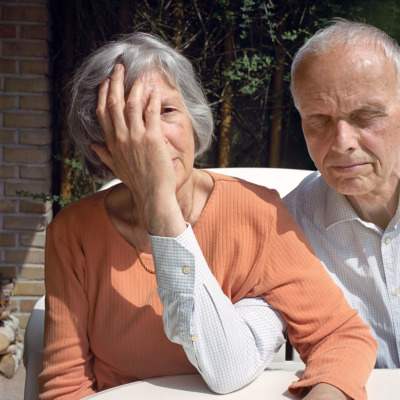 An old man closes his eyes, links arms with his wife, and covers her face.