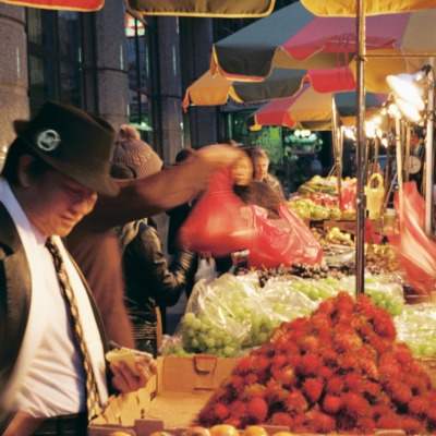 People at a local produce market are shown inspecting and purchasing vendor's goods at dusk.