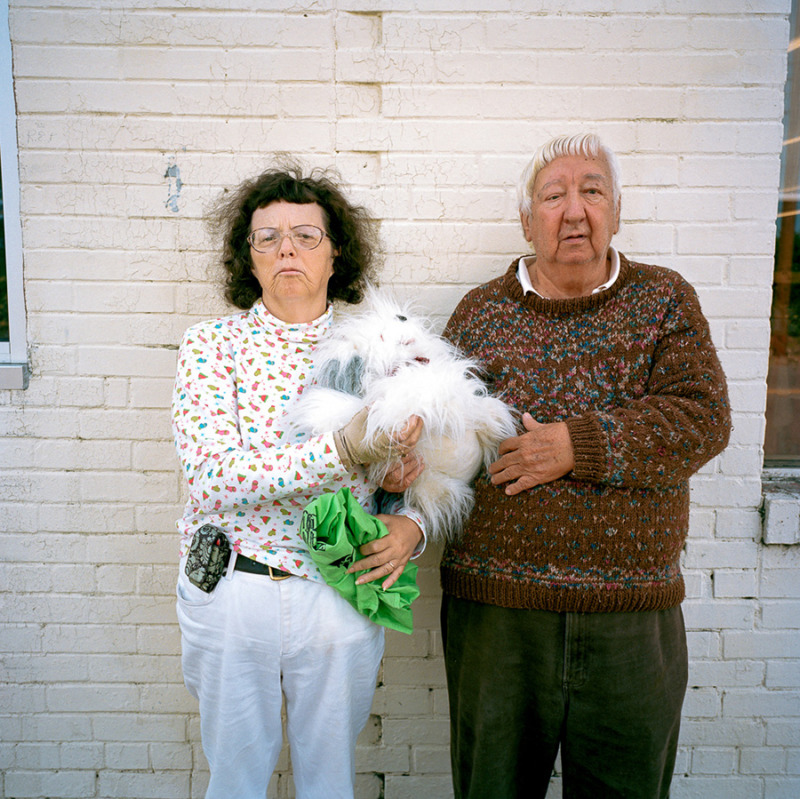 An older couple standing by a white wall, holding a small fluffy white dog