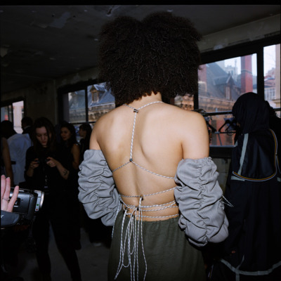 A girl with curly hair facing camera backwards wearing a top that is backless and tied together with rope, wearing grey ruched sleeves.