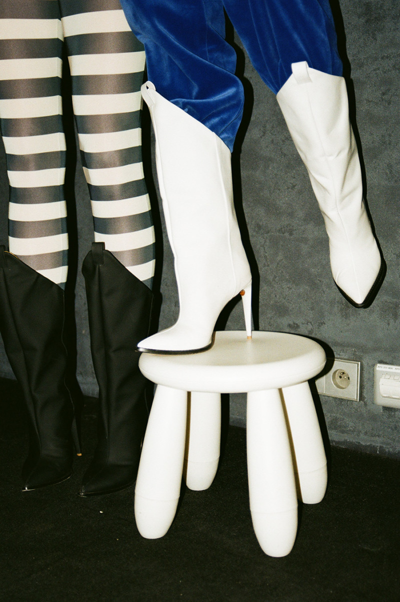 Person in white heeled tall boots and royal blue velvet trousers stands on top of a white stool, beside is a person wearing similar black boots and striped tights.