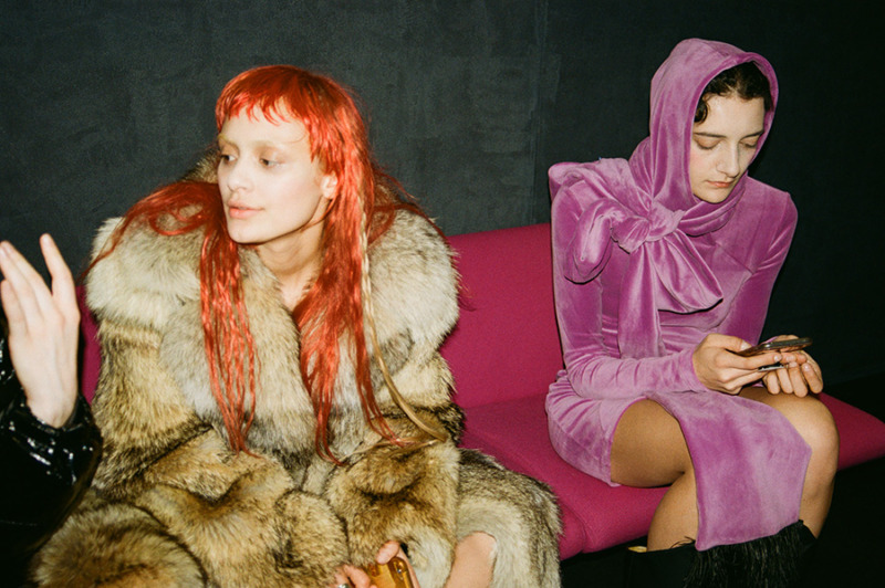 Two girls sit on a fuchsia sofa, one with red-orange hair and a thick fur coat, the other on her phone wearing a pink velvet dress and matching headscarf.