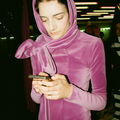 A girl is looking down at her phone wearing a pink velvet, long-sleeved dress and matching headscarf.