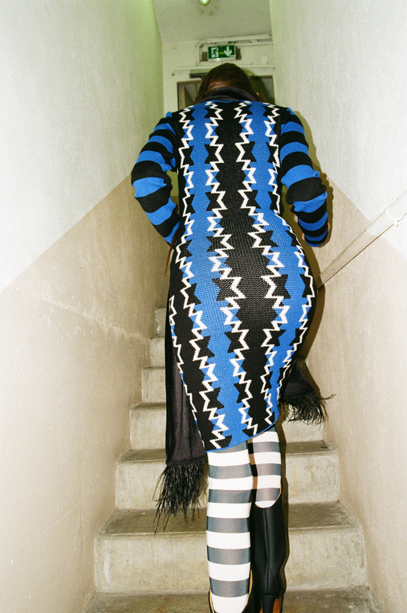 Shot of a model from behind walking up stairs wearing a geometric and striped blue and black skin-tight dress with black and white tights and black fringed scarf and black boots.
