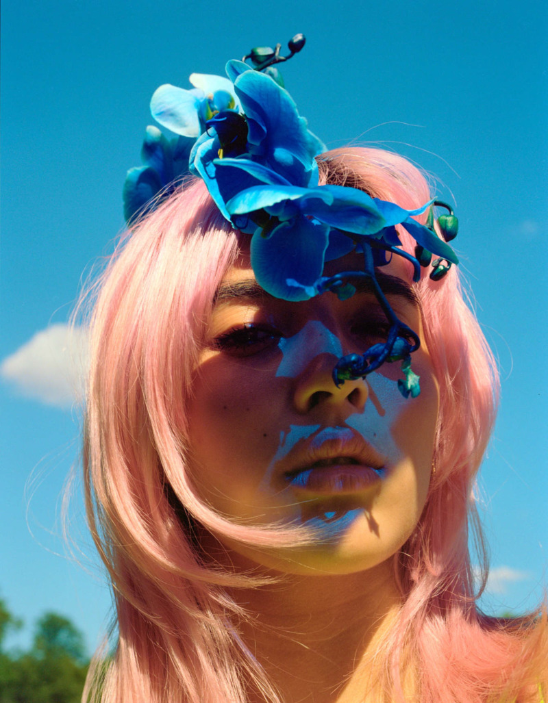 A colourful portrait of a woman's head, her pink hair contrasts with the blue flowers in her hair and the bright sky behind her.