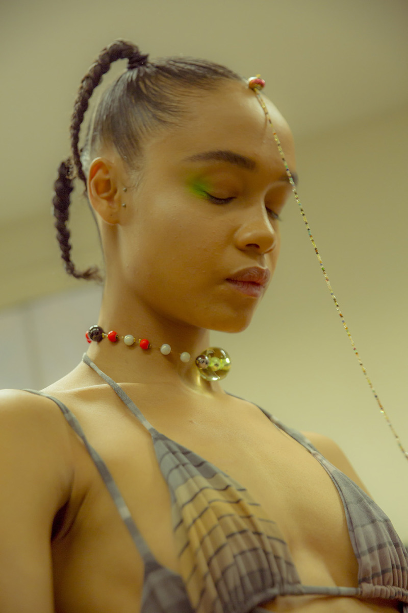A close-up of a girl with two tight ponytails in braids and a head accessory attached to hairline. Wears minimal make-up with fluorescent green eyeshadow on wings of eyes and a colourful bead necklace with a larger clear bead in centre.