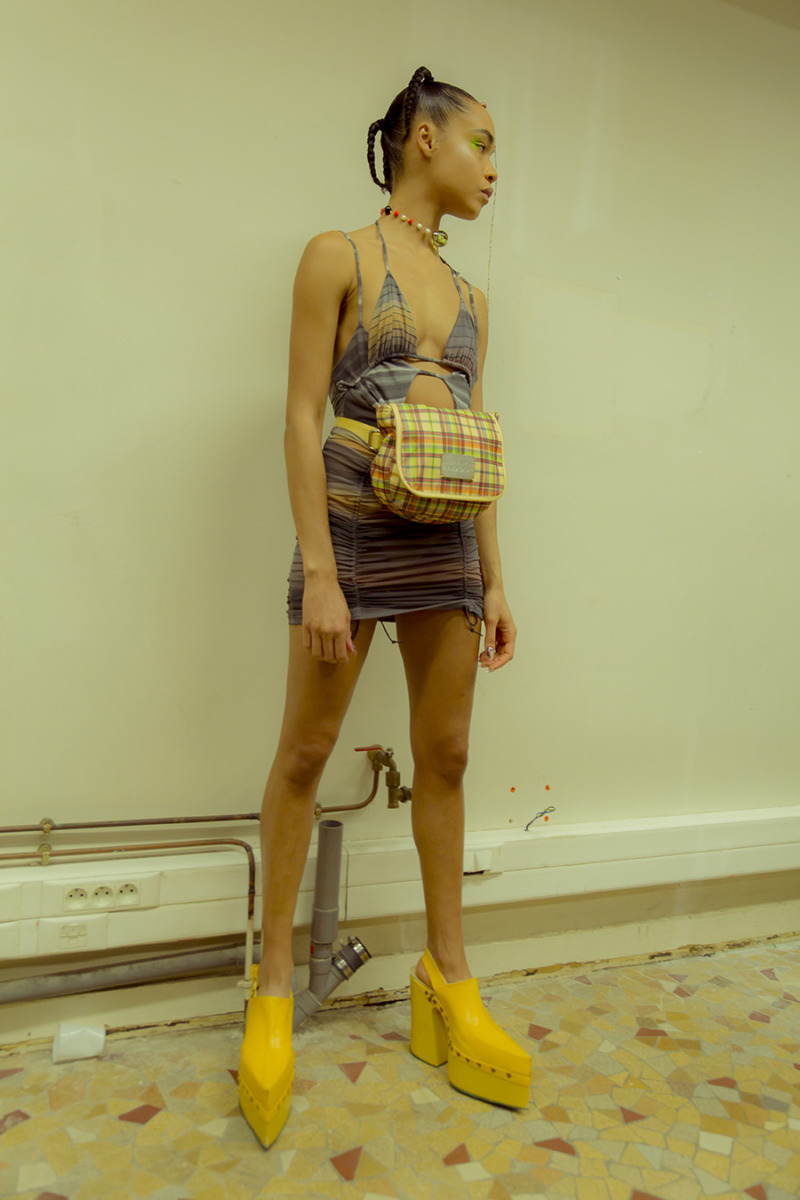A girl stands in front of a wall looking off-camera. She wears a multi-colour striped bikini-top dress with a yellow plaid fanny-pack and yellow platform ankle boots.