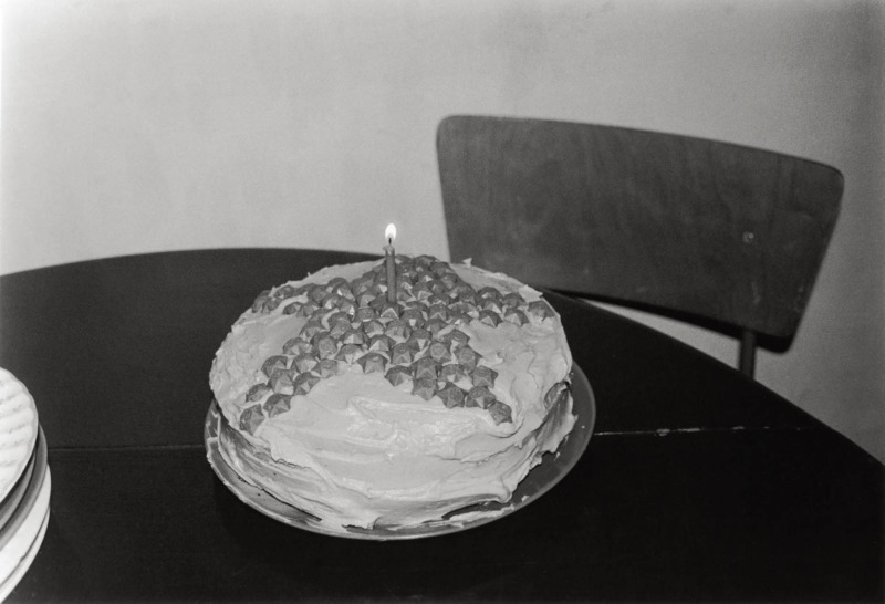 a round cake with white icing, a star made of small round chocolates on top, with a solitary lit candle, on a table top