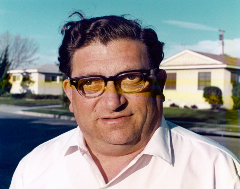A photograph of an old man with a robust face, his white shirt and thick plastic glasses might place him around the 1960s. He looks out to the camera, neither smiling nor frowning, but one eye wanders.