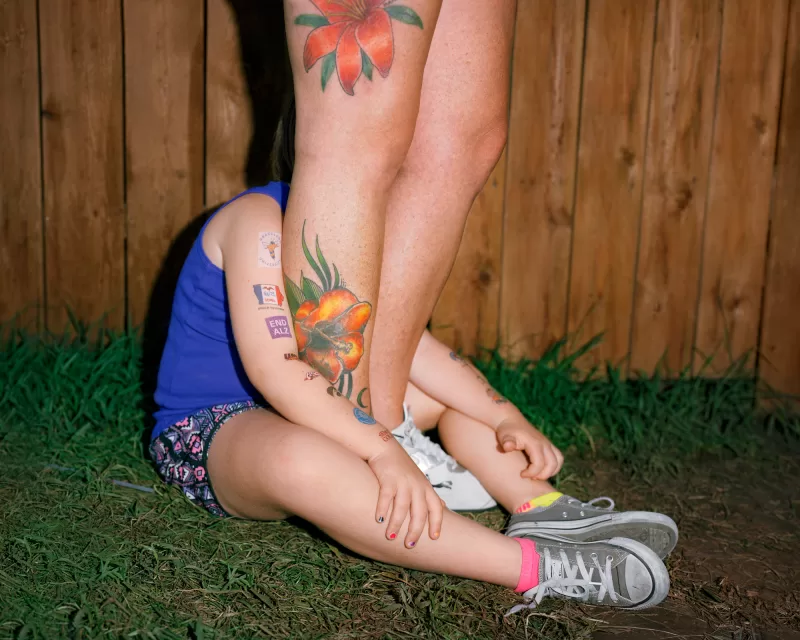 a young person sits on grass, with their face shielded by another person who stands between their legs, tattoos on their calf and thigh