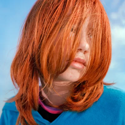 a close up portrait of a girl with bright orange hair covering her face and wearing a blue sweater, looks over her left shoulder