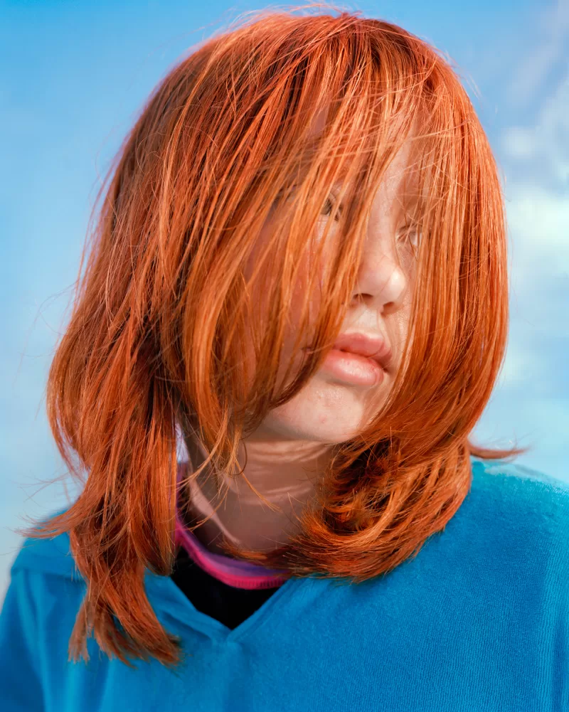 A close up portrait of a girl with bright orange hair covering her face and wearing a blue sweater, looks over her left shoulder.