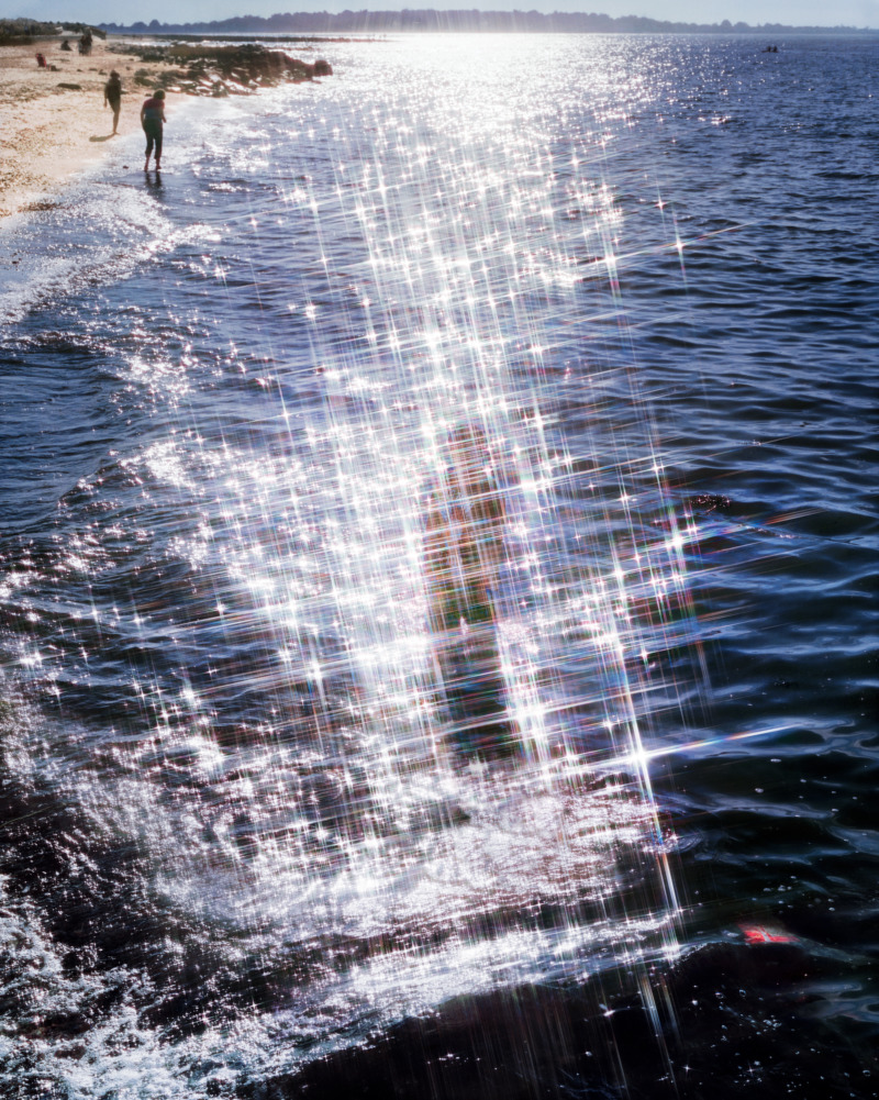 A person standing in the water along the shoreline, sun is glistening in the water