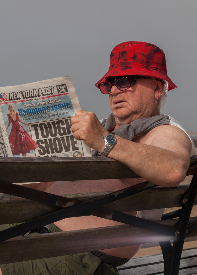 An old white man in a bucket hat reads a tabloid paper on a bench.