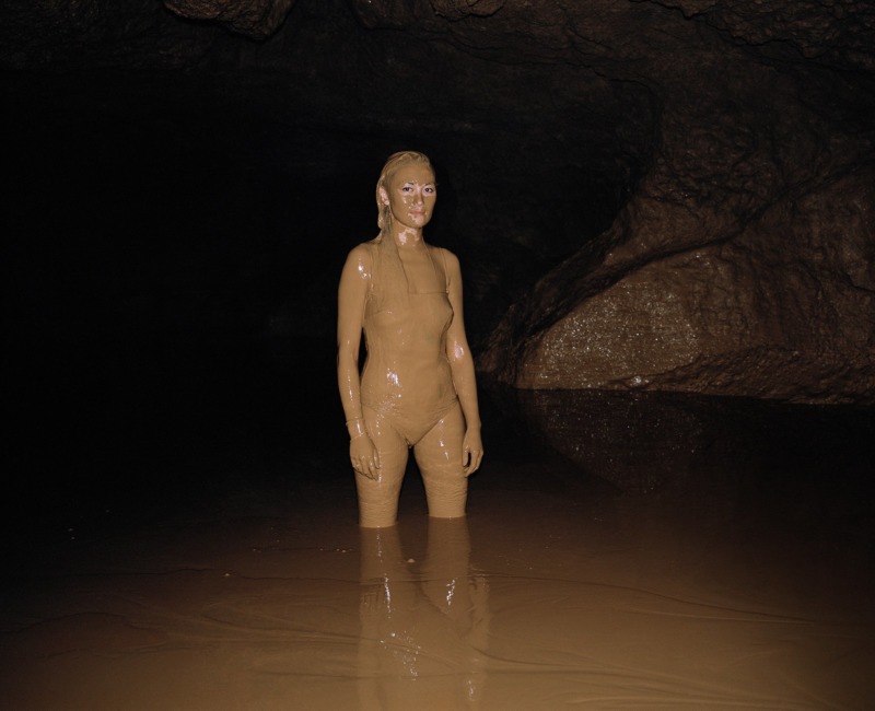 A woman, coated in mud, stands alone in a cave.