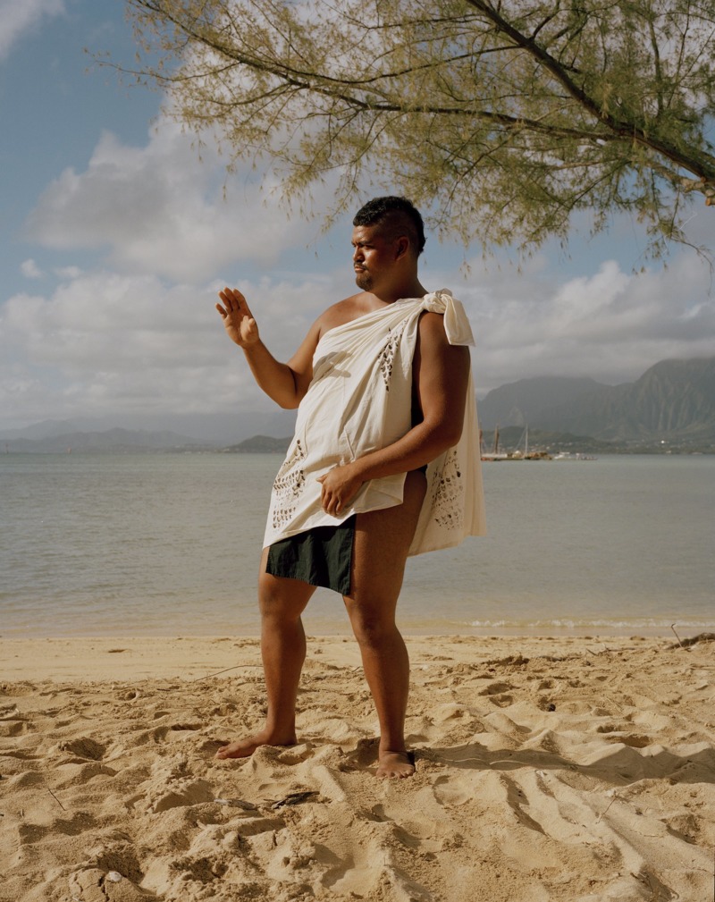 A man in traditional Hawai'ian dress stands on a beach, gesturing off-camera.