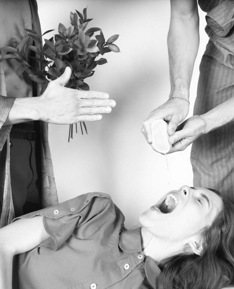 In what looks like some strange ritual a woman lies back and drinks the juice of a lemon being squeezed into her mouth by unseen man while another man holds over her a bouquet of leaves.