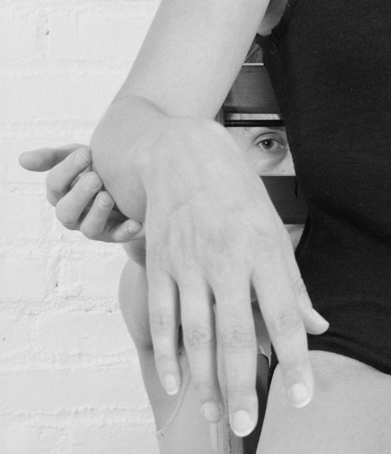 A foreshortened hand is extended out towards the camera, another anonymous hand holds their elbow. In the gap between arm and body is the eye of a person, otherwise hidden.