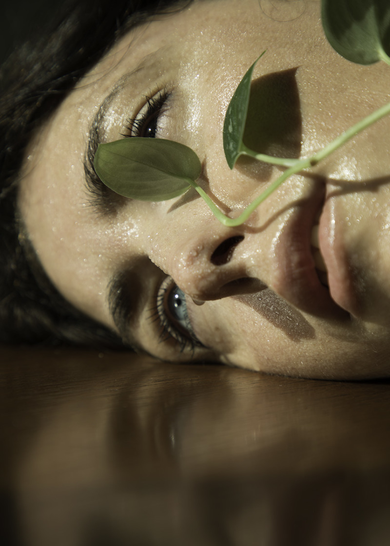 A woman's warm, damp face lies against a metal surface, a few leaves gently brushing her lips.