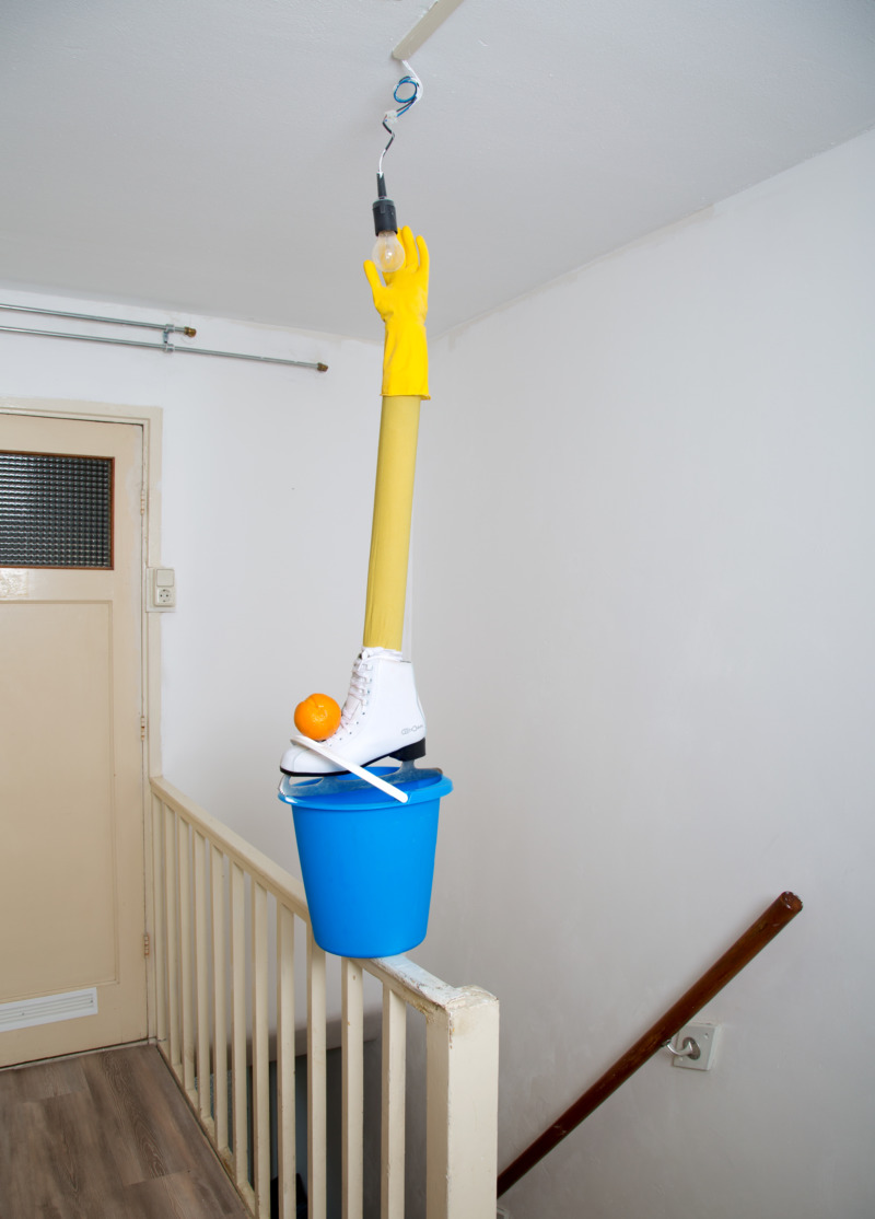 A marigold glove stands on a pipe, a shoe, and a bucket, to reach a lightbulb.
