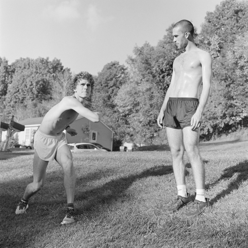 Two shirtless young men stand on the manicured lawn of an American suburb. They appear like ancient athletes, the first contorts his body round to hurl a frisbee, the second stands tall, watching.