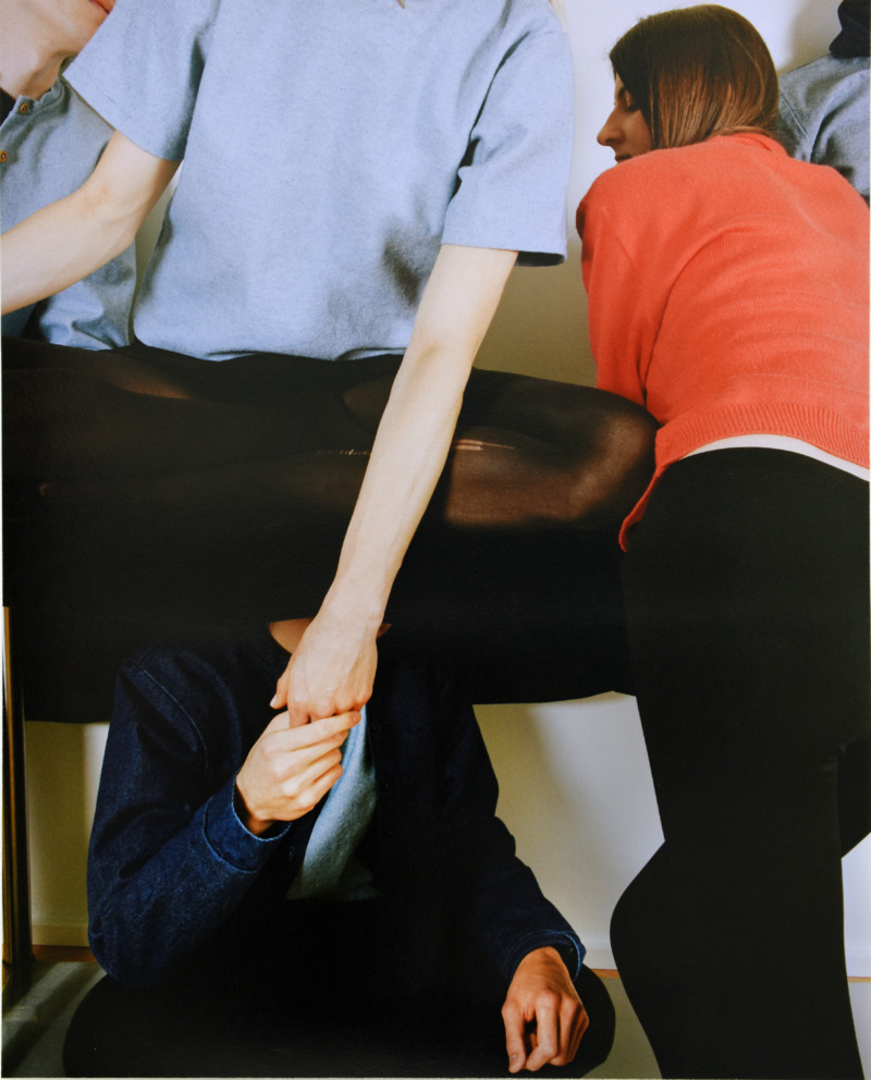 Three people sit above, around, and underneath a table, their colourful jumpers contrasting with each other, their three bodies forming an almost abstract composition.