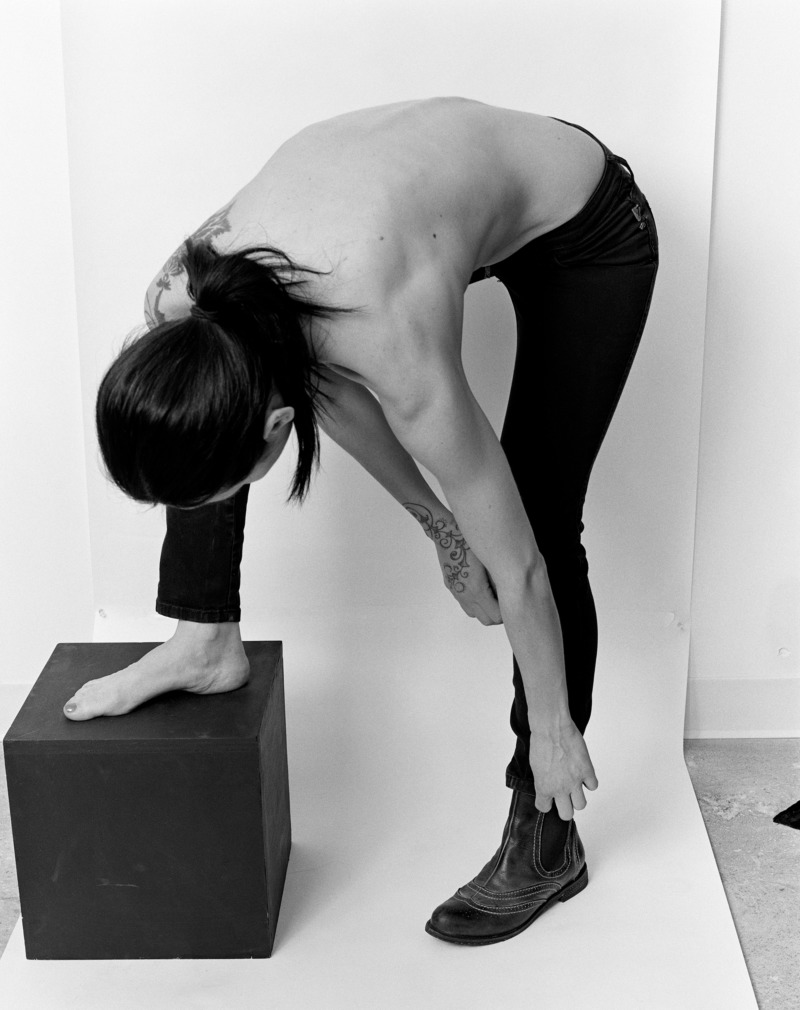 A shirtless person stood in a photography studio takes a moment to bend down and adjust their sock.