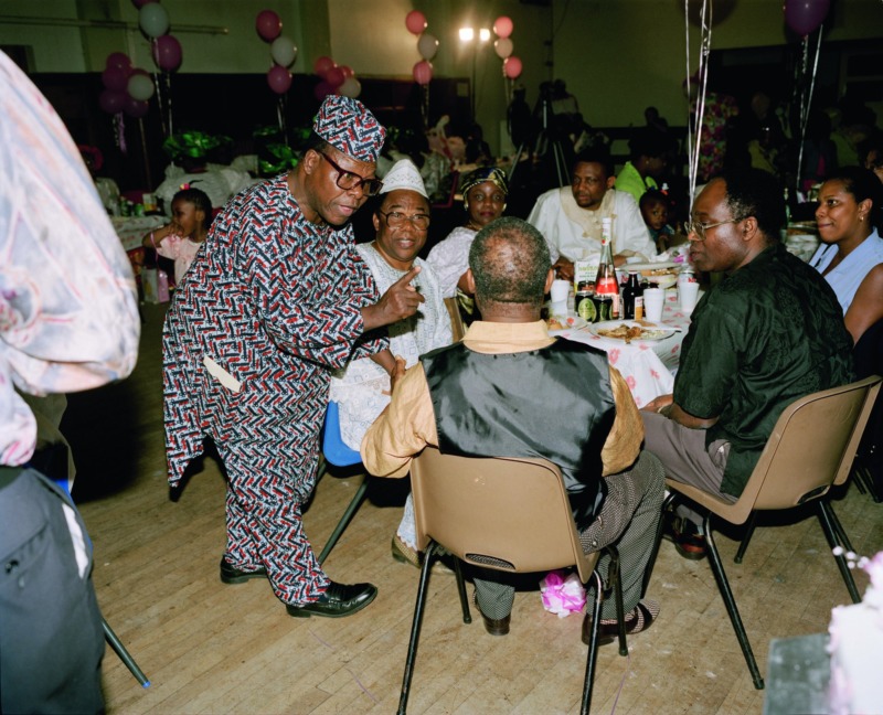 A group of old Nigerian men sit around a table in a function room, engaged in an animated conversation. Plates of food and empty bottles of Supermalt and Guinness dot the table.