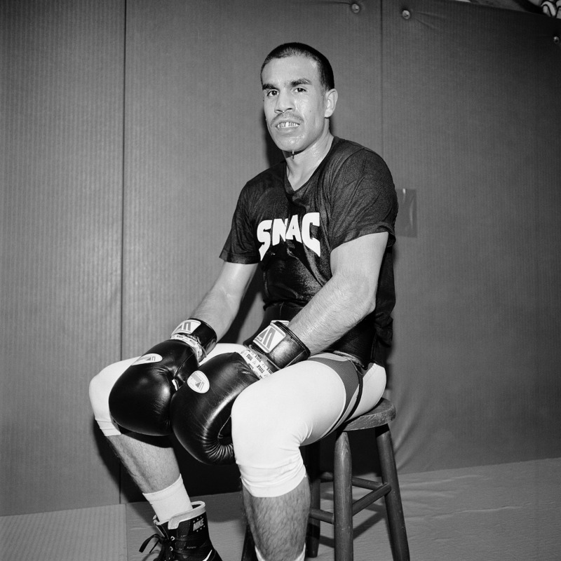 An older boxer, his gloves on and head uncovered, sits on a wooden chair in a bare room.