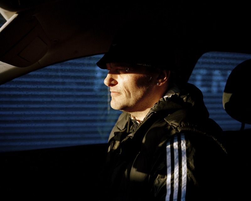 A moody, darkly lit portrait of a man in a tracksuit sat in a car.