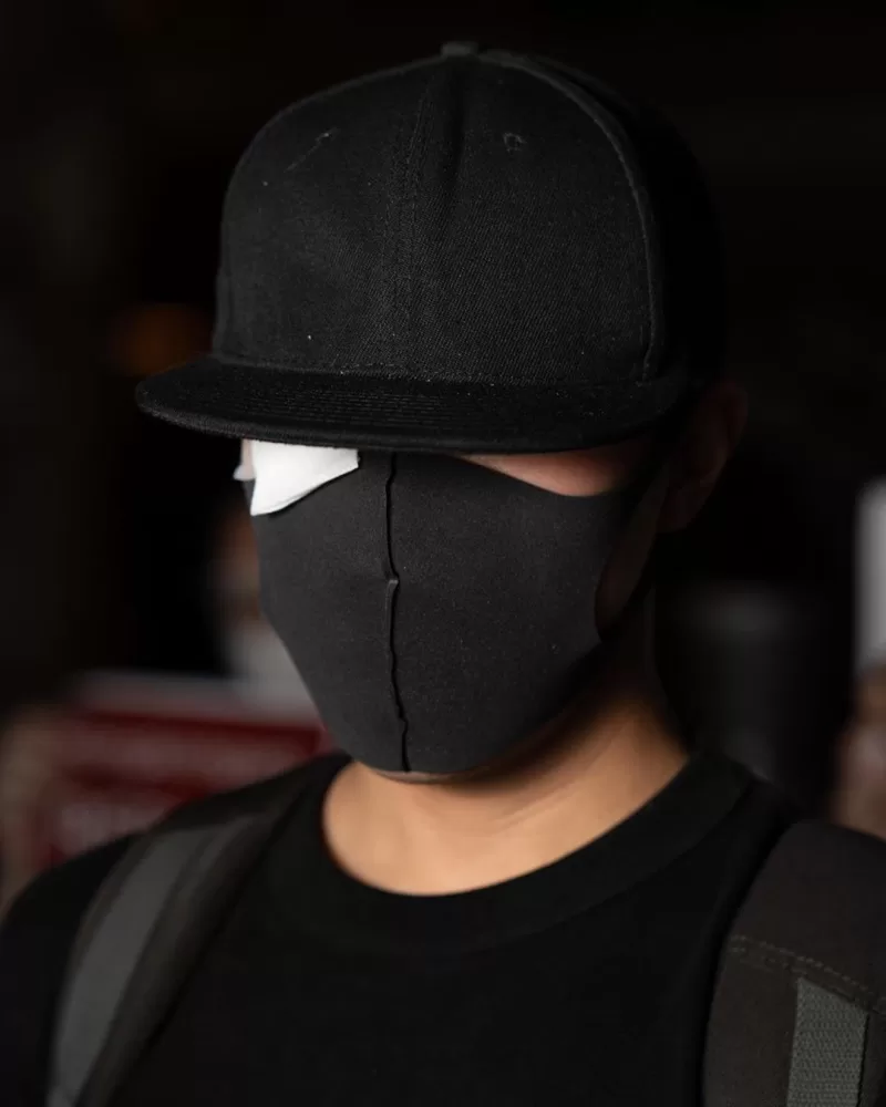 A young person in a black t shirt covers their face with a black mask and a black cap.