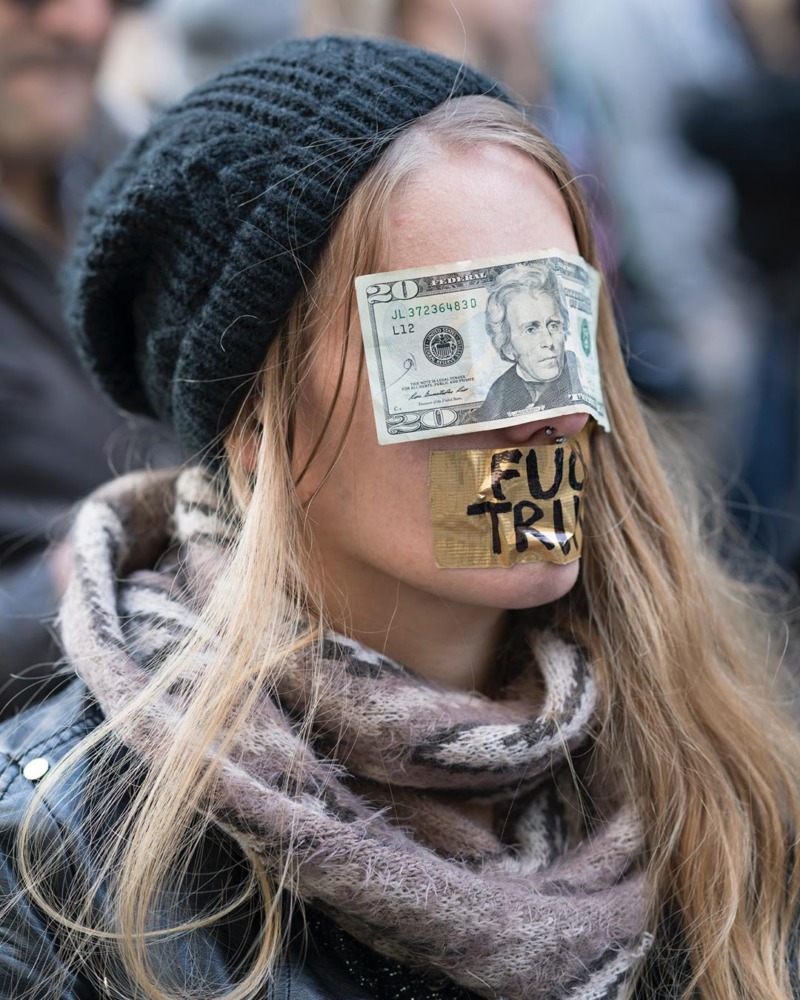 A young woman has her face covered with a dollar bill and a sticker saying 'FUCK TRUMP'.