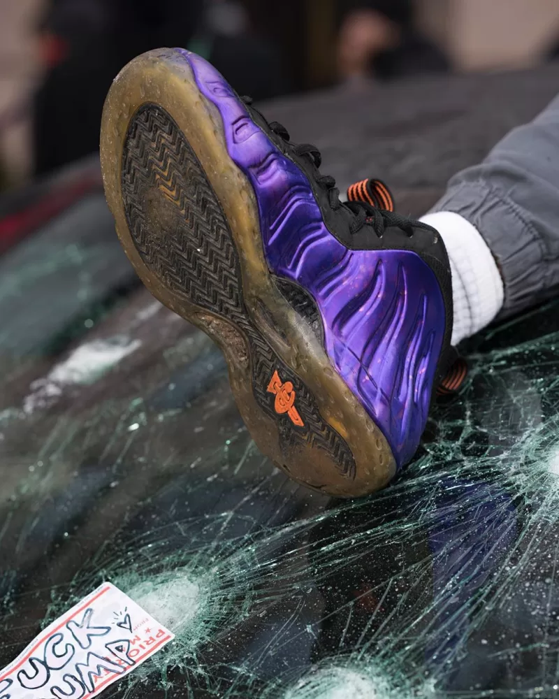 A foot rests on the shattered windowscreen of a car, a sticker in the bottom left says 'UCK -UMP'