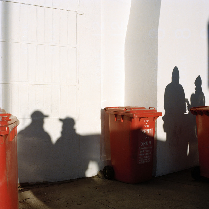 A group of chatting men cast their shadows over a while wall and a row of red bins.