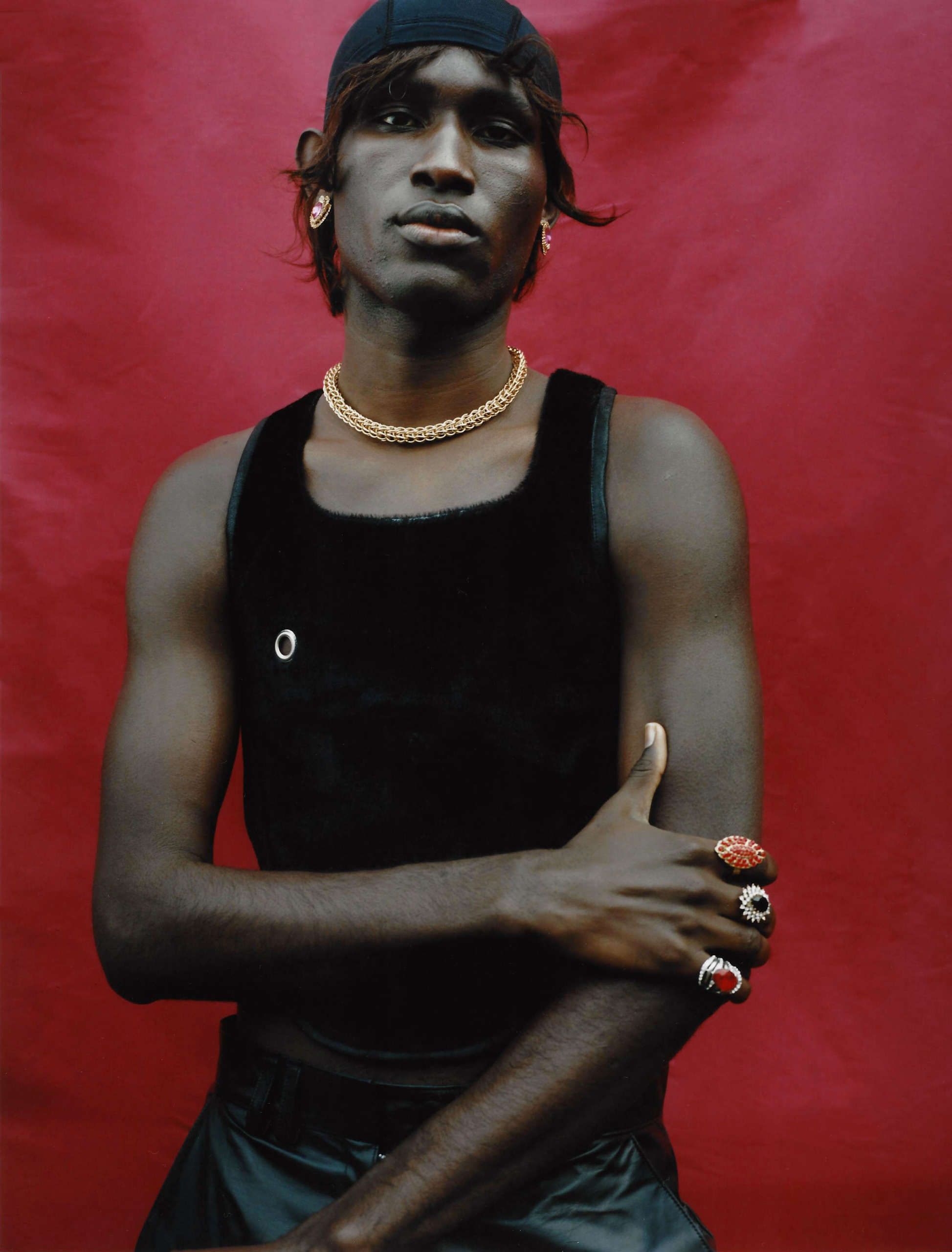 A man wearing a black tank top, black pants, and three red, black, and silver rings poses in front of a red background.