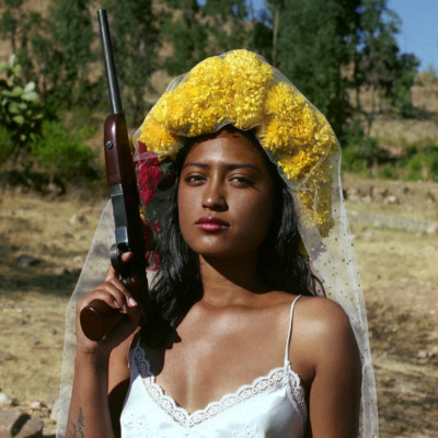 A young woman wearing a wedding dress and carrying a gun stares at the viewer.