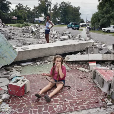 A young girl holding a snake sits in the ruins of a demolished home. Nobody seems concerned by the scene.